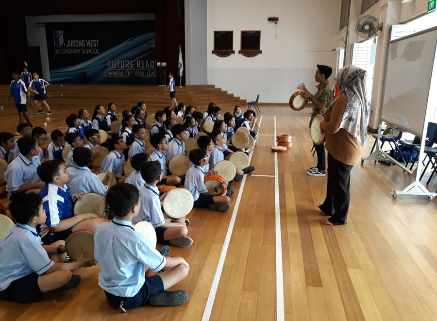 Students in uniform sit on a gym floor holding frame drums while two instructors demonstrate at Jurong West Secondary School.